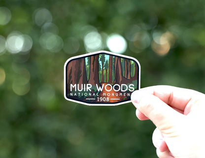A hand holds the Muir Woods National Monument sticker with its redwood trunks against blurred green foliage.