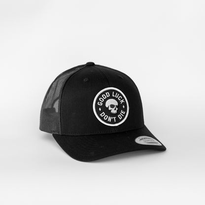 An all black Good Luck Don't Die trucker hat shows its skull patch clearly against the dark mesh and brim.