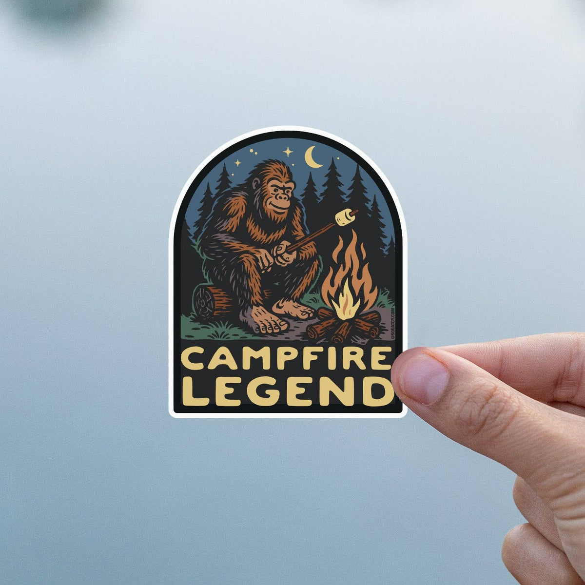 A thumb holds the Campfire Legend sticker flat against soft blue glass, moon and stars clear above Bigfoot.