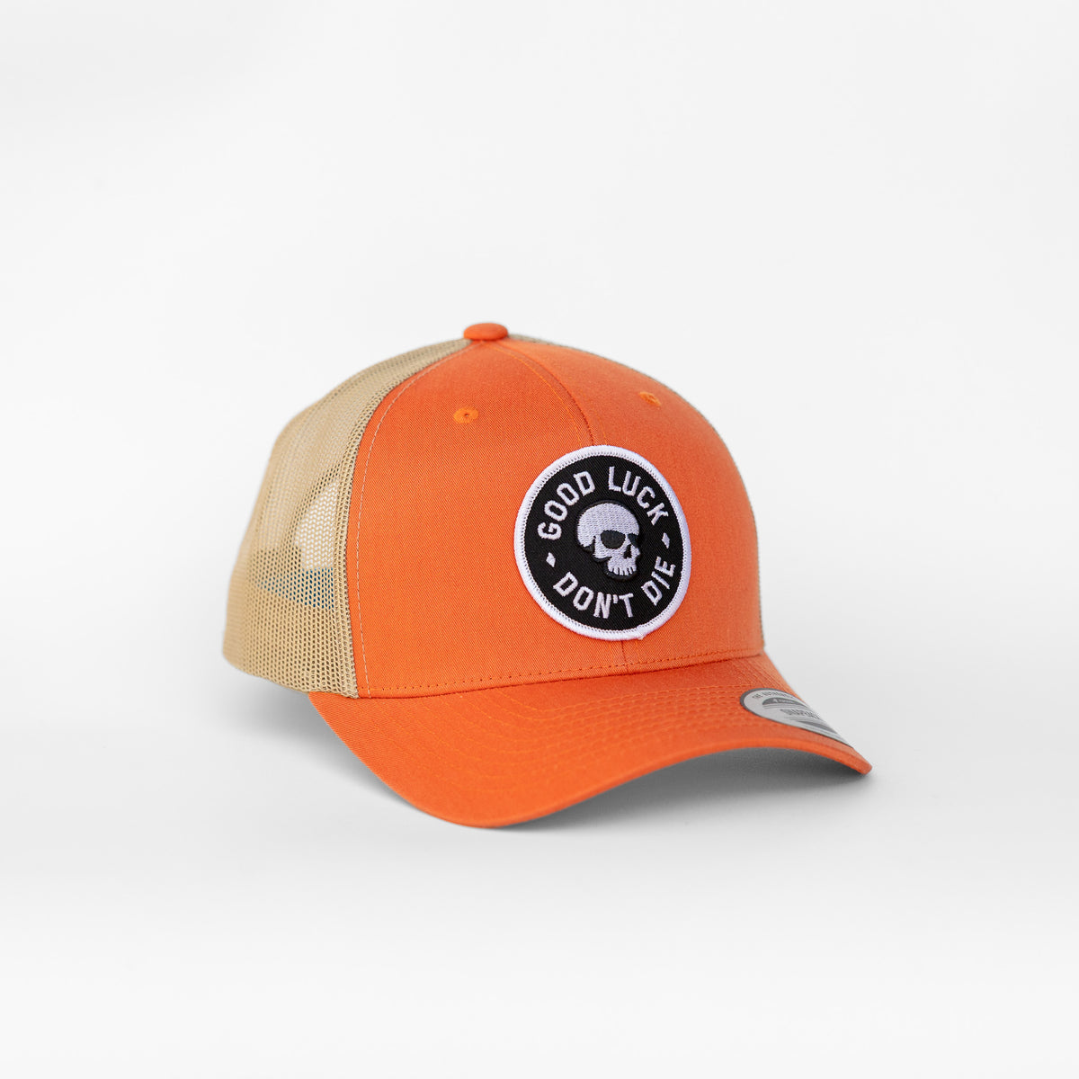 An orange front panel on the Good Luck Don't Die trucker hat carries a round skull patch above tan mesh sides.