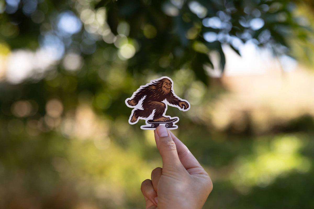 Backlit leaves fill the background as fingers hold up the small Later Skater Bigfoot sticker.