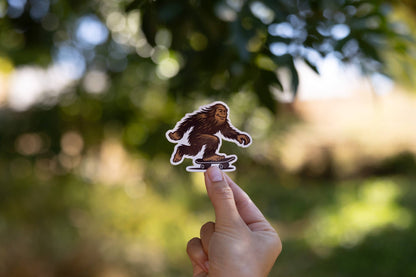 Backlit leaves fill the background as fingers hold up the small Later Skater Bigfoot sticker.