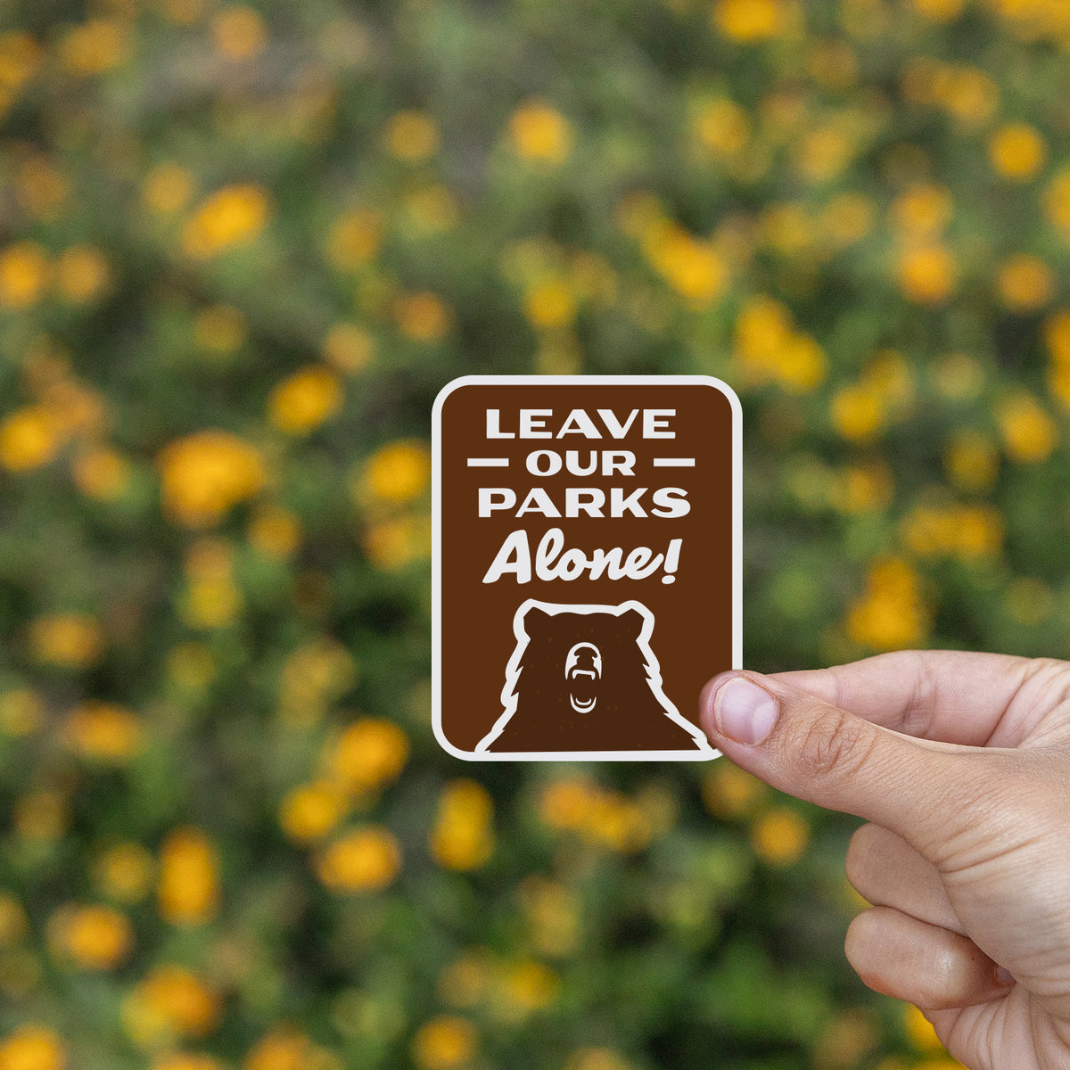 Held between two fingers, the Leave Our Parks Alone sticker stands out against blurred yellow wildflowers.