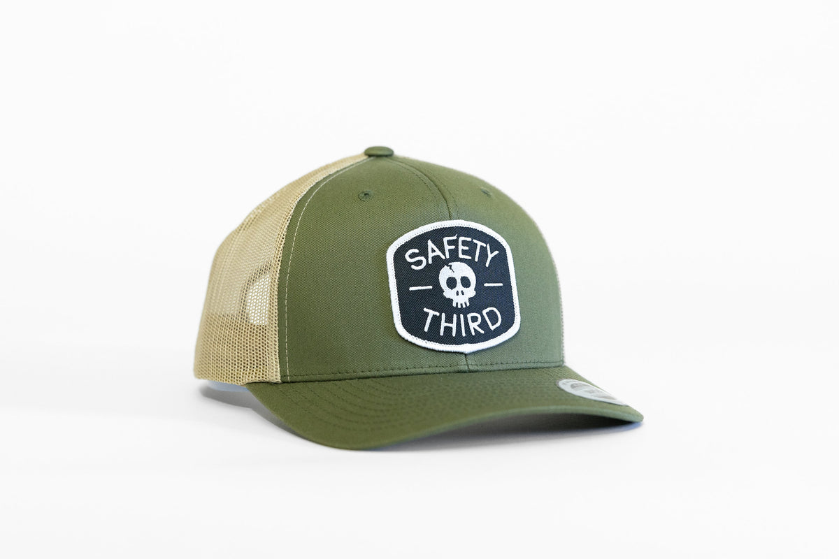 The olive Safety Third trucker hat sits with tan mesh sides and the white bordered skull patch facing front.