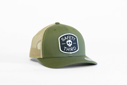 The olive Safety Third trucker hat sits with tan mesh sides and the white bordered skull patch facing front.