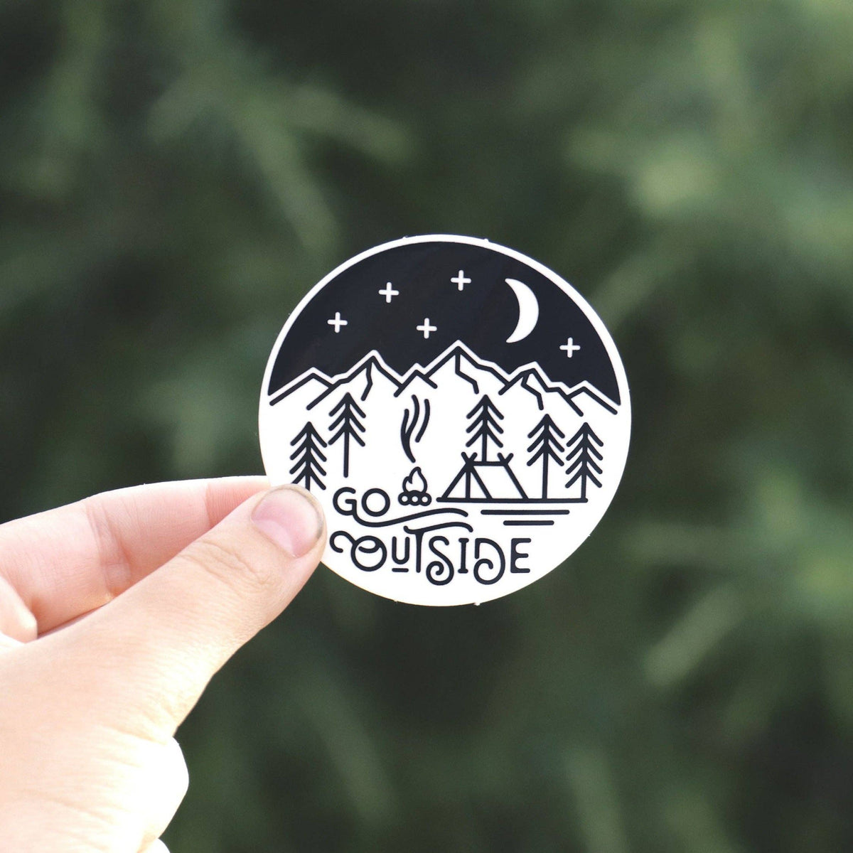 Fingers pinch the round Go Outside sticker close to the lens, its campfire and crescent moon clear against green pines.