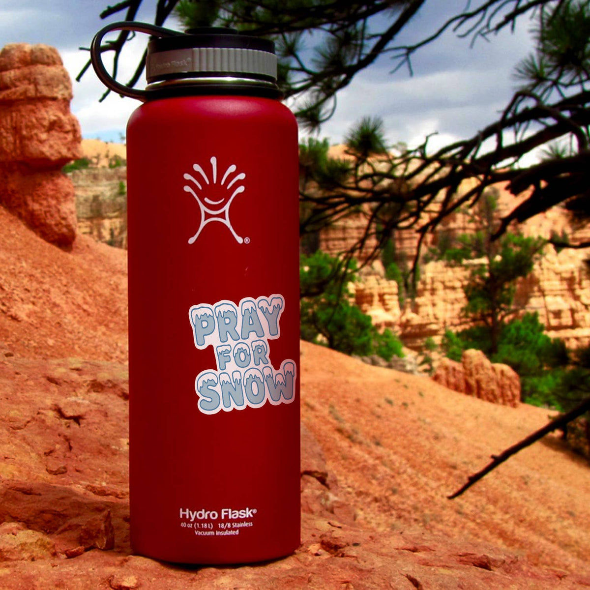 A red Hydro Flask wears the Pray For Snow sticker on red rock with hoodoos and pines behind.