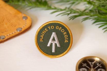 Beside a leather sheath and a brass compass, the AT Maine to Georgia patch catches soft light.