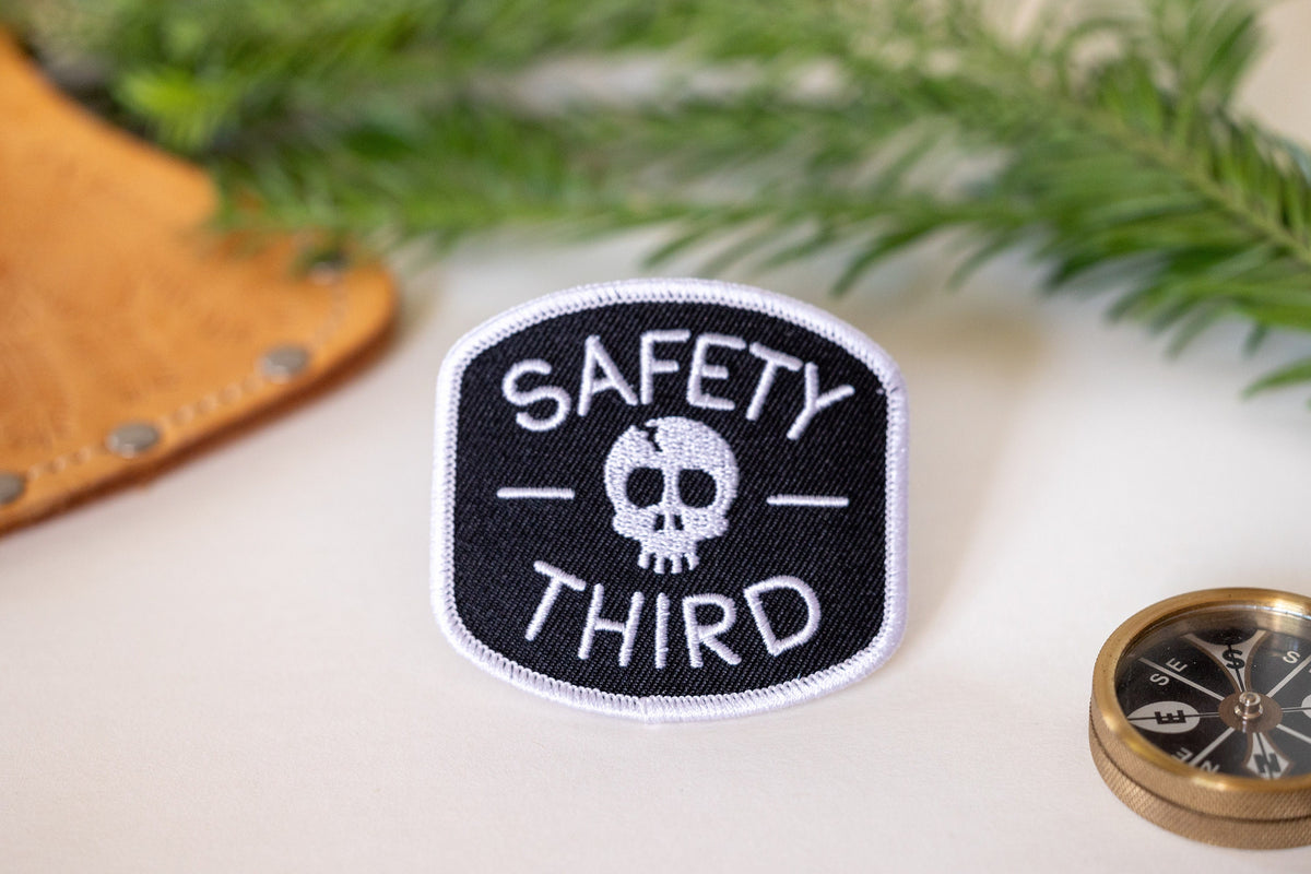 Beside a brass compass and a pine sprig, the Safety Third patch sits on a cream tabletop with leather nearby.