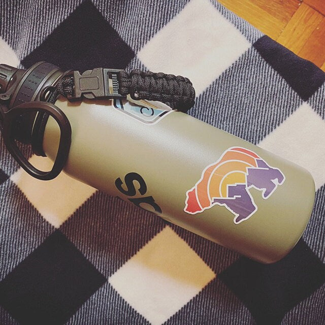 An olive water bottle with a paracord strap wears the Adventure Grizzly Bear sticker on a plaid blanket.