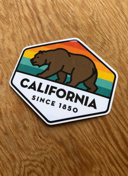 Resting flat on light oak grain, the hexagon California State Bear sticker reads SINCE 1850.