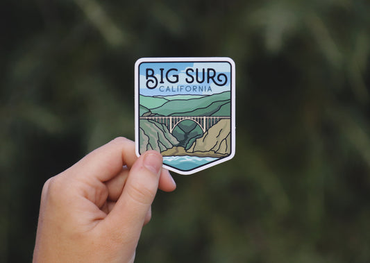 Held further back against dark evergreens, the Big Sur California sticker shows the full shield shape and lettering.
