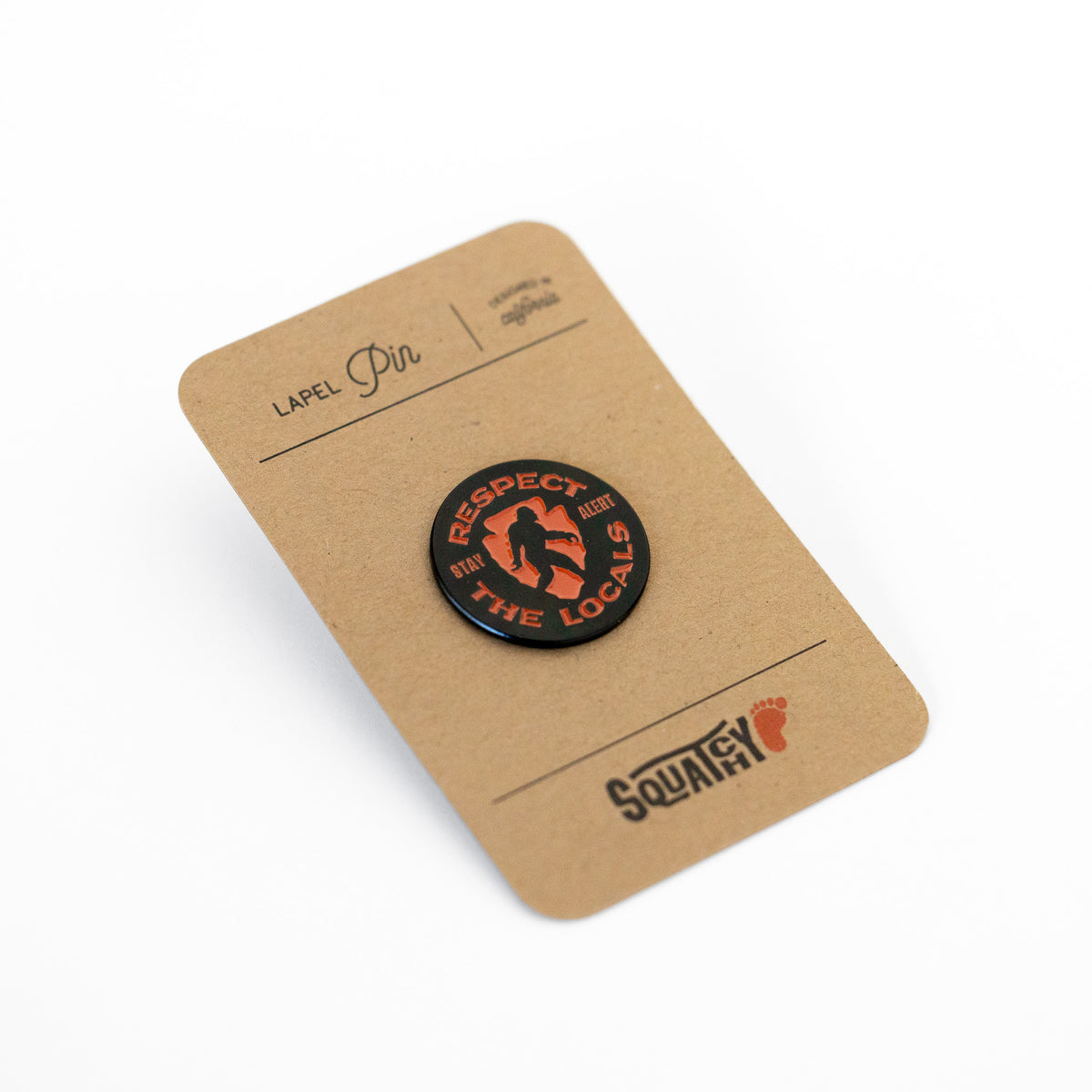 Still on its kraft backing card labeled Lapel Pin, the Respect The Locals pin sits above the Squatchy footprint stamp.