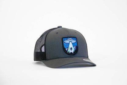 Charcoal and black Bigfoot UFO Conspiracy trucker hat angled on white, patch showing a saucer beaming up a sasquatch.