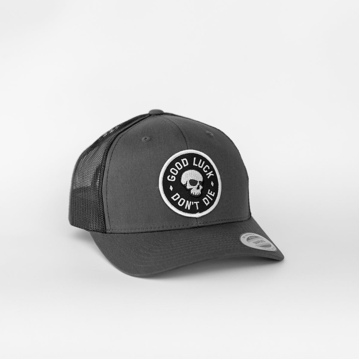A charcoal grey crown on the Good Luck Don't Die trucker hat frames the round skull patch above its curved brim.