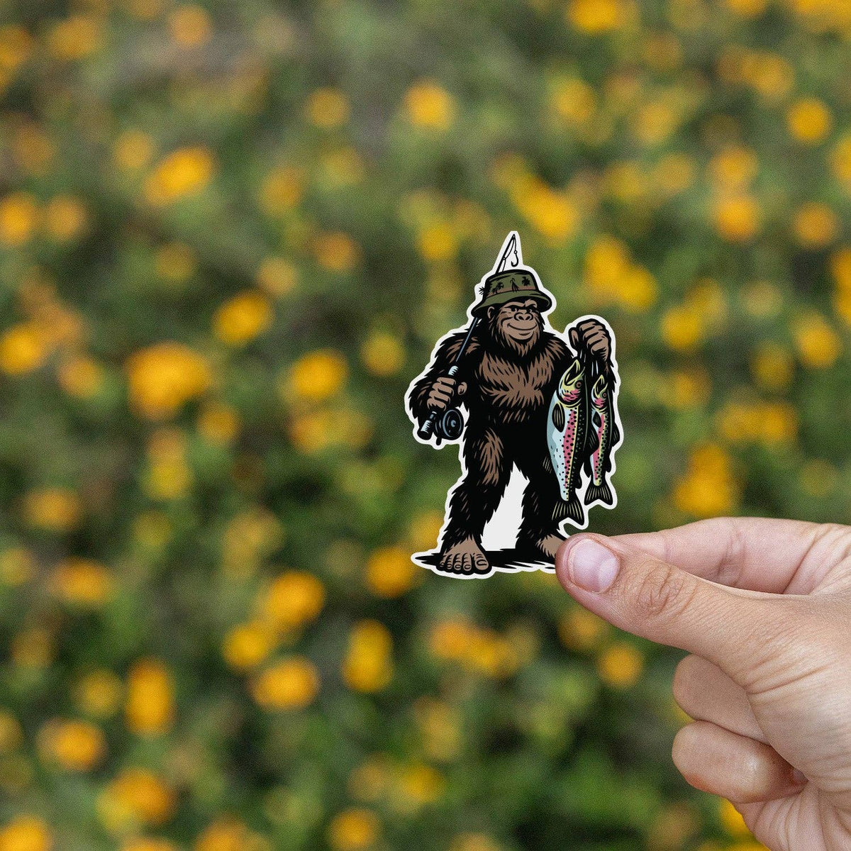 Pinched between thumb and finger, the Bigfoot Fishing sticker floats over a field of yellow blooms.