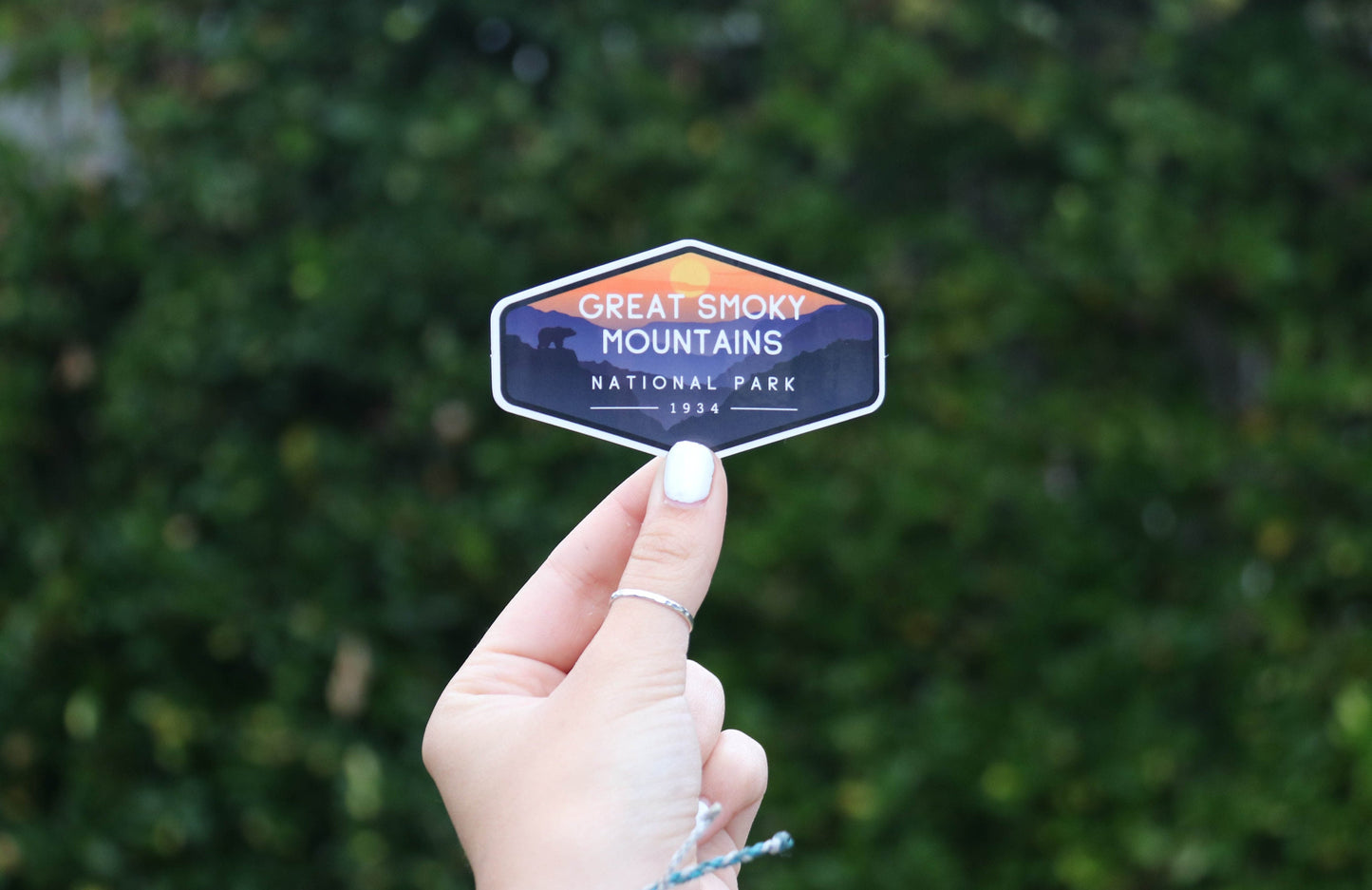 Pulled back from the leaves, a hand with white nail polish holds the small Smoky Mountains National Park sticker.