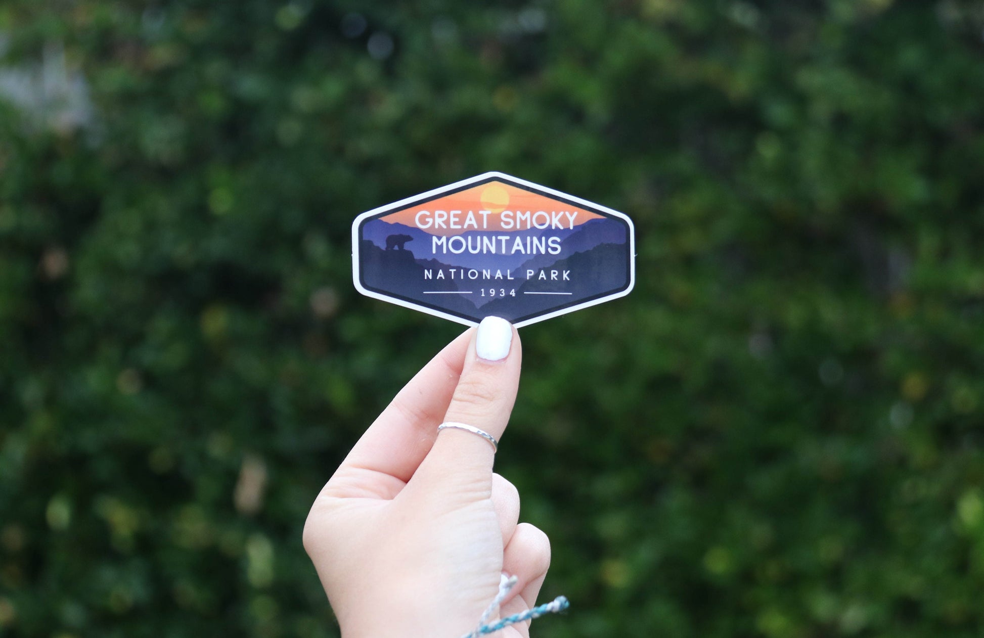 Pulled back from the leaves, a hand with white nail polish holds the small Smoky Mountains National Park sticker.