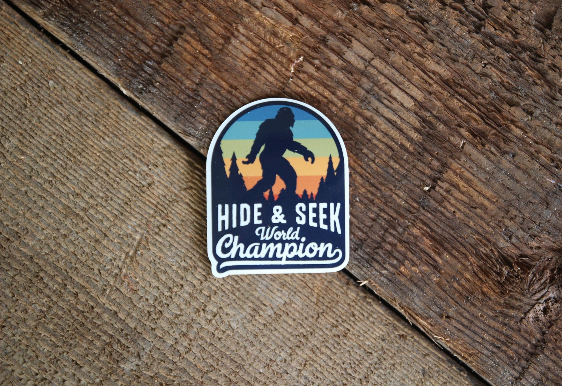 Resting on weathered brown barn wood, the Bigfoot Hide & Seek World Champion sticker lies flat.