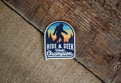Resting on weathered brown barn wood, the Bigfoot Hide & Seek World Champion sticker lies flat.