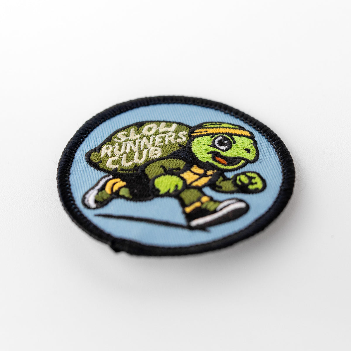 Seen at an angle with its edge curling slightly, the Slow Runners Club patch displays the embroidered turtle mid stride.