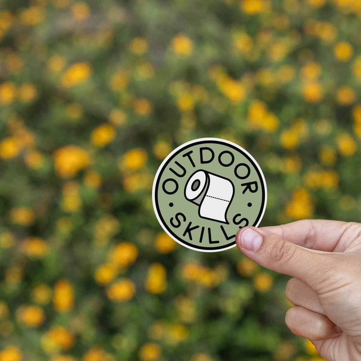 Yellow wildflowers blur behind the Outdoor Skills sticker pinched between finger and thumb.