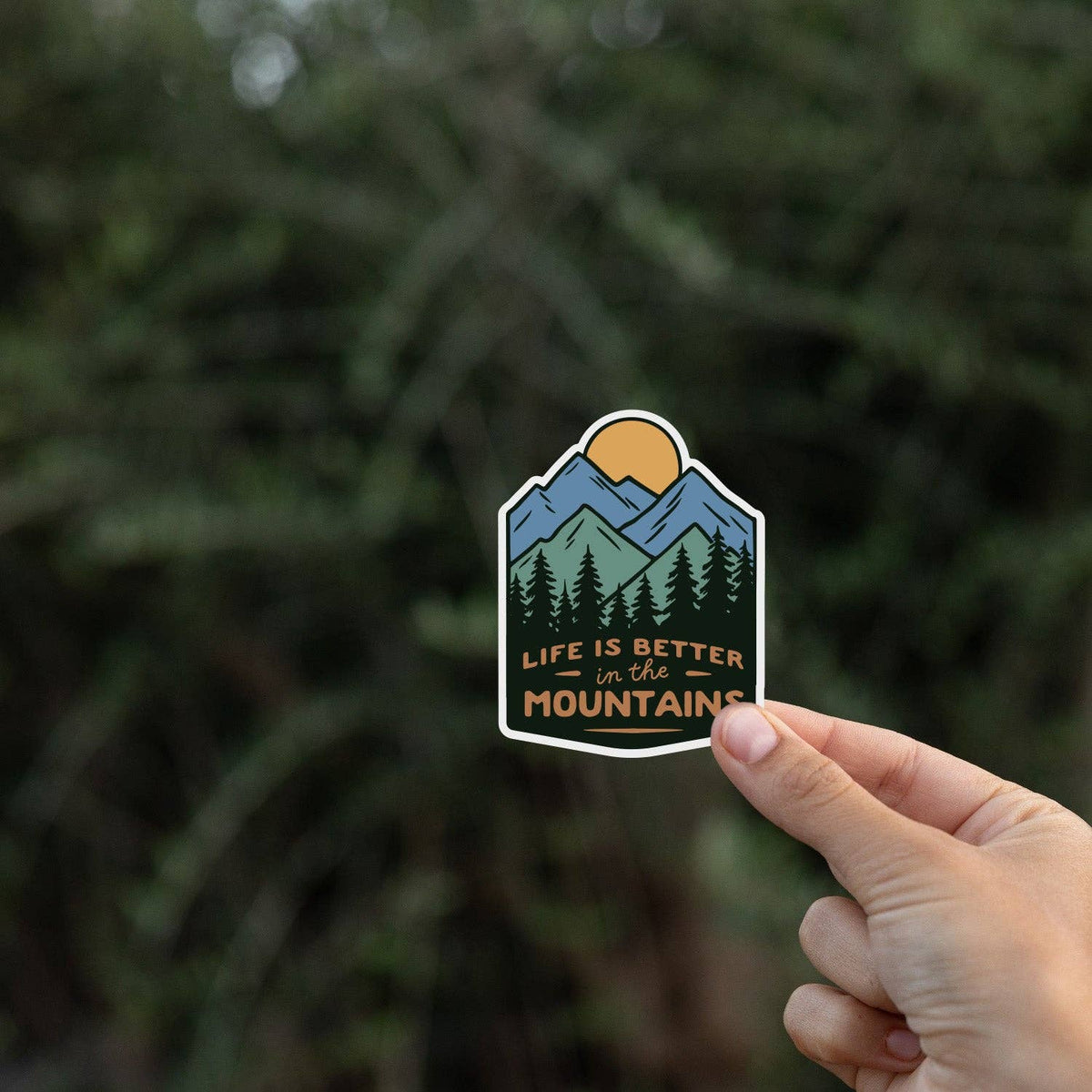 A hand holds the Life Is Better In The Mountains sticker against a blurred wall of dark evergreens.