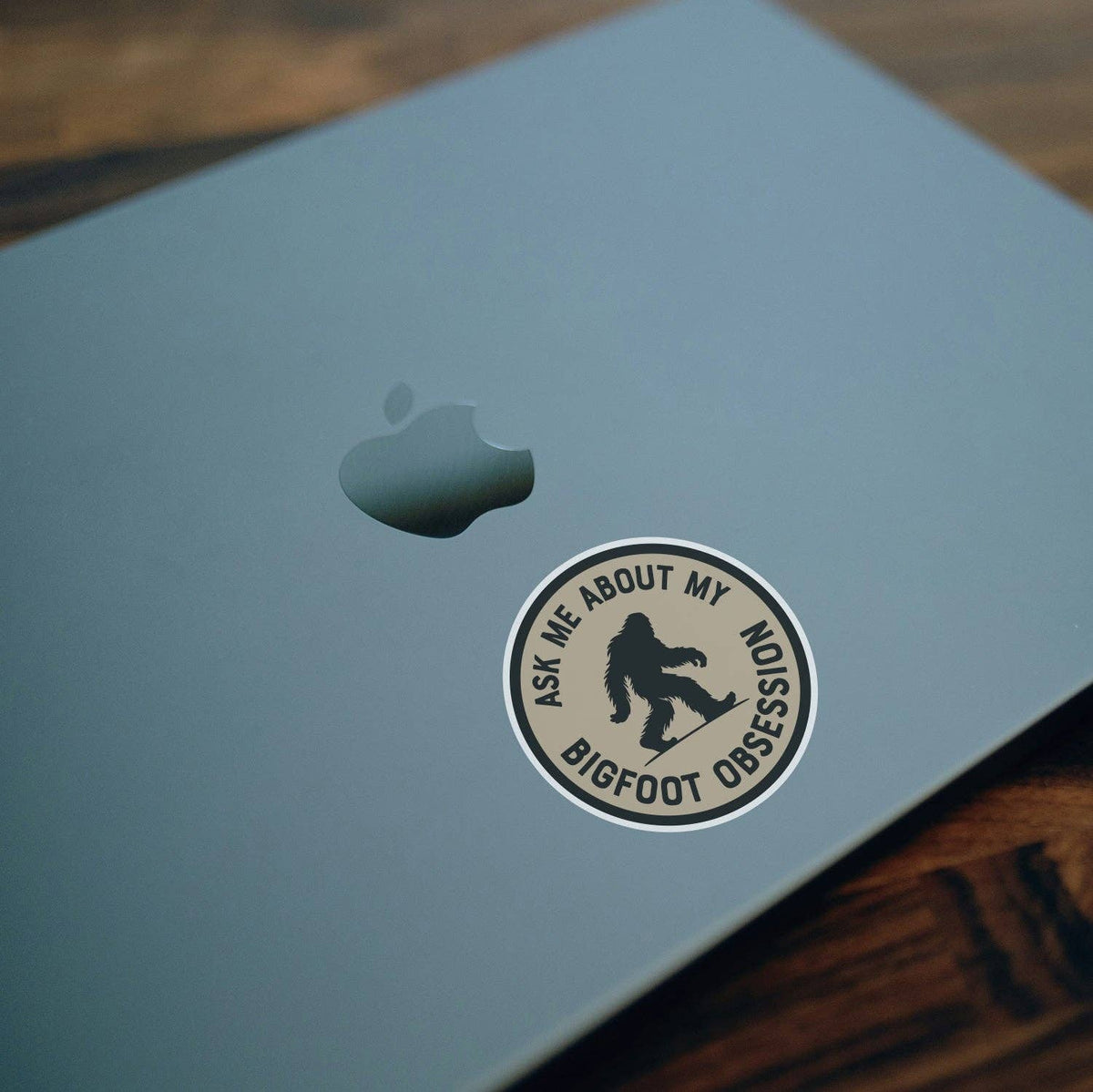 On the lid of a pale blue laptop, the Ask Me About My Bigfoot Obsession sticker sits below the apple logo.
