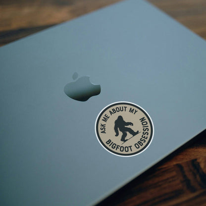 On the lid of a pale blue laptop, the Ask Me About My Bigfoot Obsession sticker sits below the apple logo.