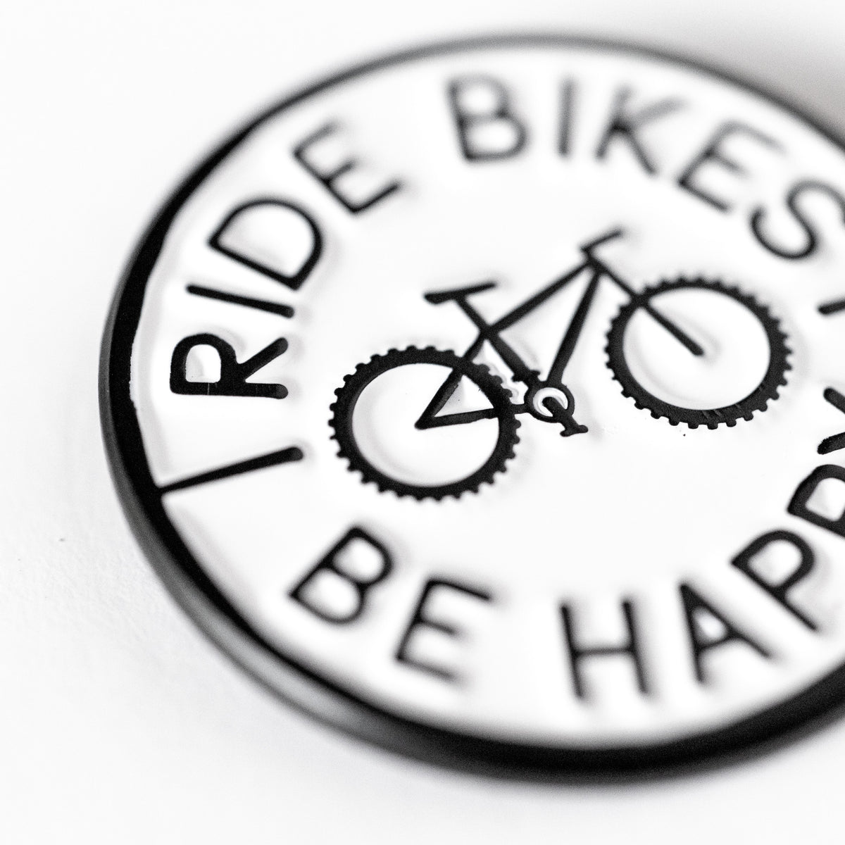 Shot at a close angle, the Ride Bikes Be Happy pin shows raised lettering and a bicycle in sharp relief.