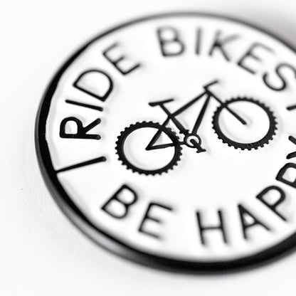 Shot at a close angle, the Ride Bikes Be Happy pin shows raised lettering and a bicycle in sharp relief.