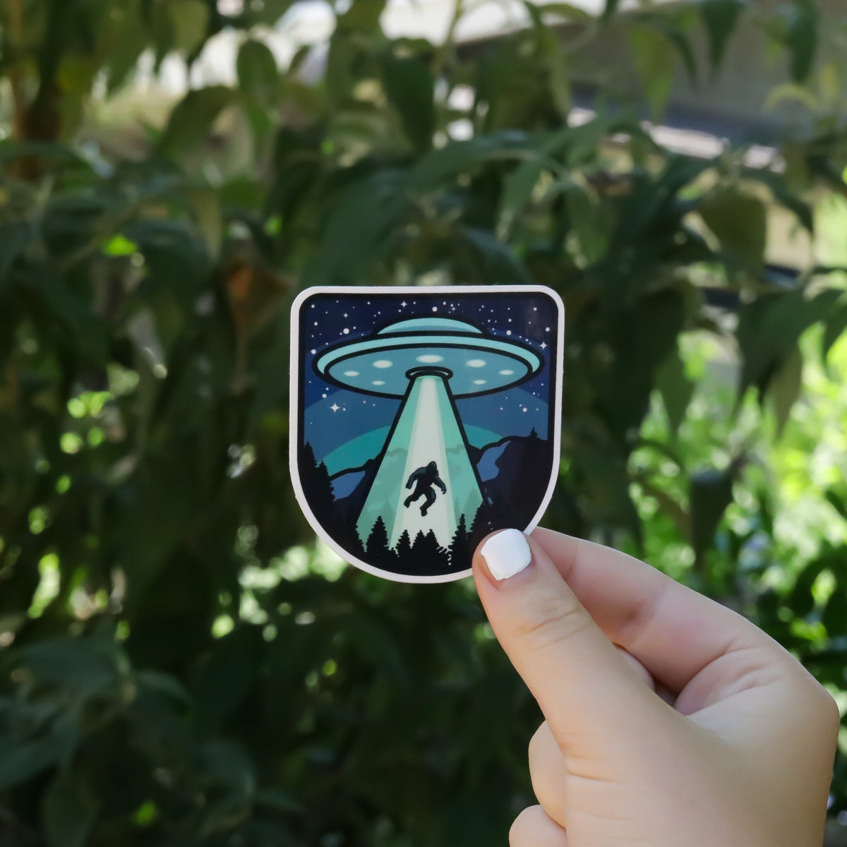 White painted nails hold the Bigfoot UFO Conspiracy sticker in sunlight before a leafy hedge.