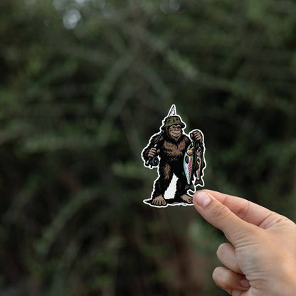 Held by one corner, the Bigfoot Fishing sticker stands against a shadowy stand of evergreen branches.