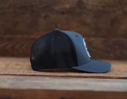 Side profile on rough planks, the charcoal Safety Third trucker hat shows its mesh panel and curved brim.