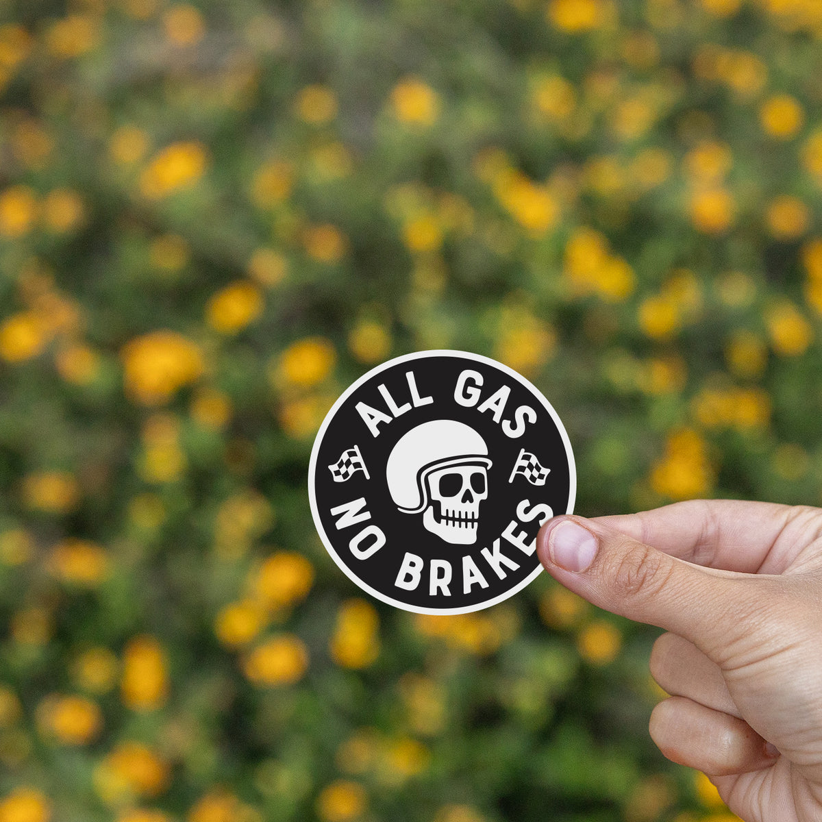 Fingers pinch the All Gas No Brakes sticker in front of a blurred field of small yellow flowers.