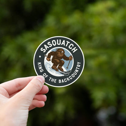 Thumb and finger grip the Sasquatch King of the Backcountry sticker with green summer leaves far behind.