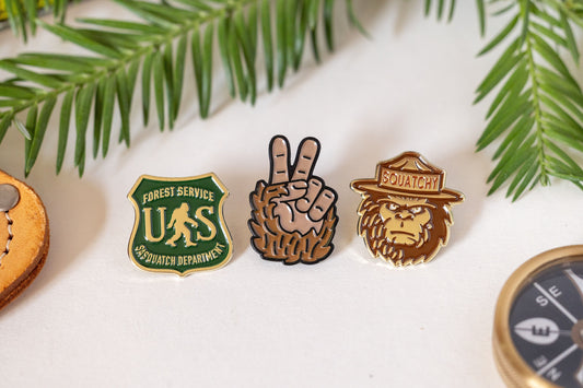 Three enamel pins in a row, Sasquatch Department shield, peace hand and Squatchy ranger, under pine branches.