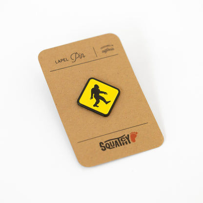 Mounted on a kraft paper lapel pin card, the Bigfoot Crossing pin sits above the Squatchy footprint logo.
