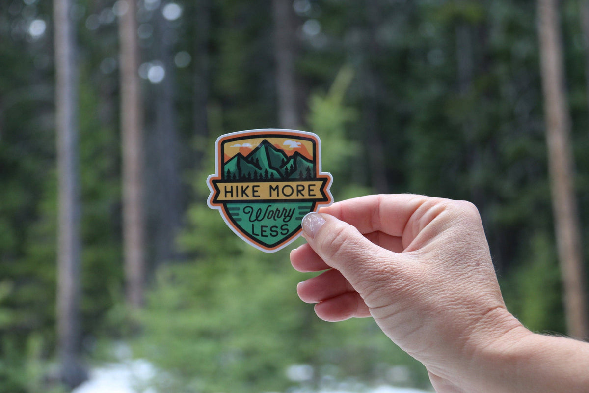 Deep in a pine forest, fingers grip the Hike More Worry Less sticker with its mountain and sunrise art.