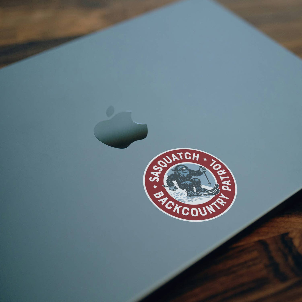 On the lid of a pale grey laptop beside the Apple logo, the Sasquatch Backcountry Patrol sticker rests at an angle.