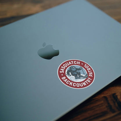On the lid of a pale grey laptop beside the Apple logo, the Sasquatch Backcountry Patrol sticker rests at an angle.