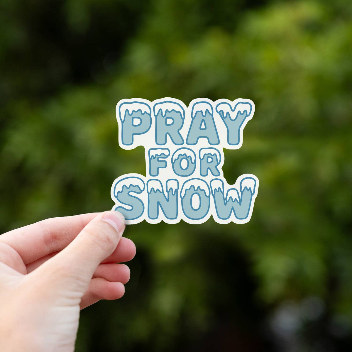 Held higher against the same leafy blur, the plain Pray For Snow sticker shows its icy drip lettering.