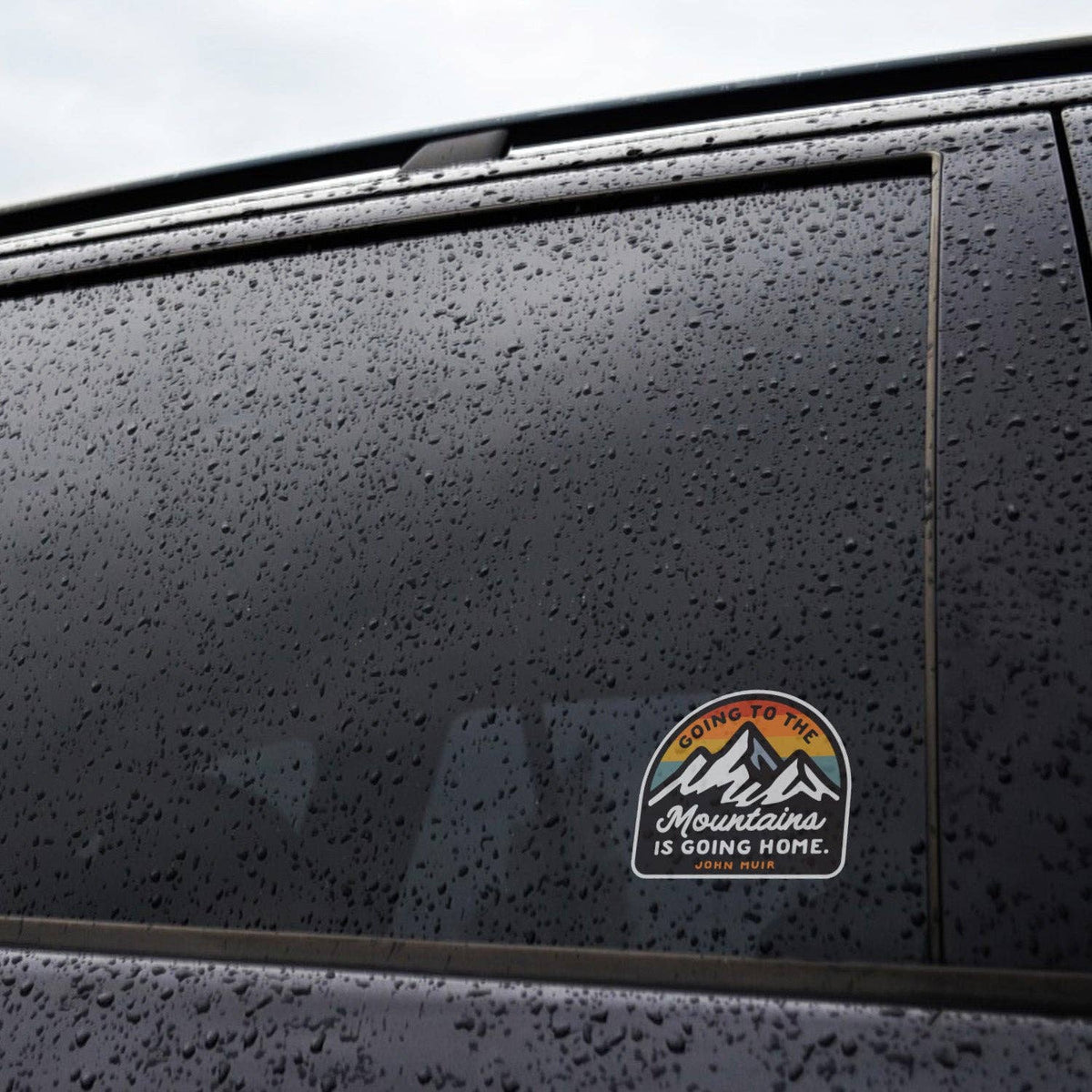 Rain beads on a dark car window where the Going to the Mountains is Going Home sticker is applied.