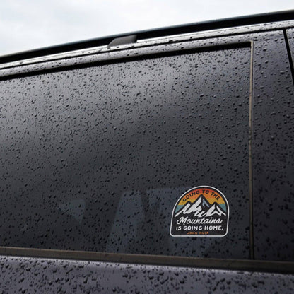 Rain beads on a dark car window where the Going to the Mountains is Going Home sticker is applied.
