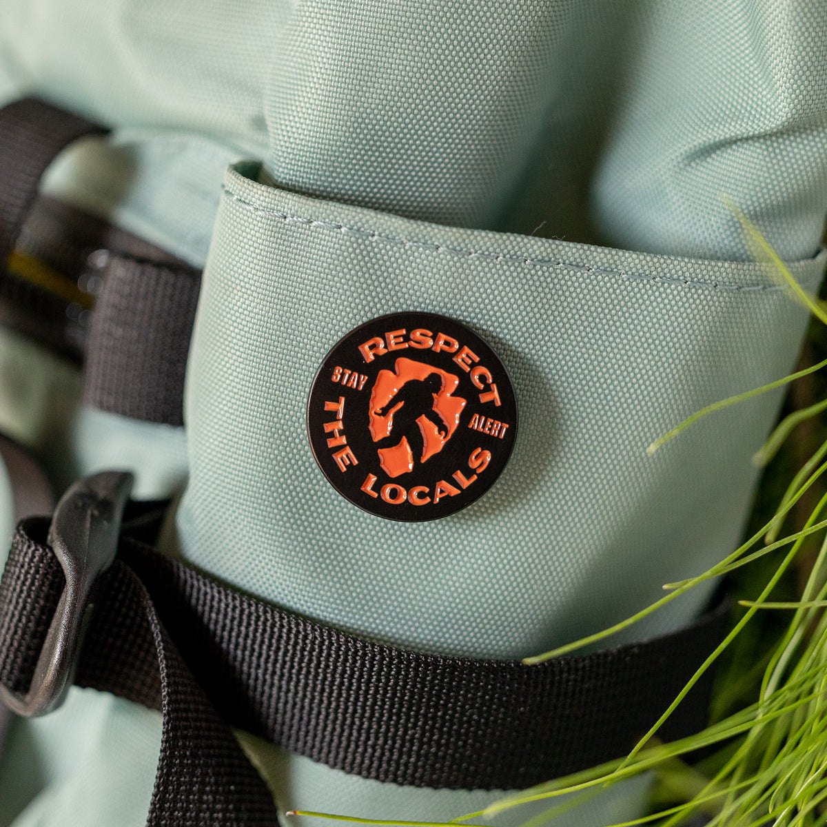 Clipped to a sage green pack pocket, the Respect The Locals pin sits beside blades of grass and a buckled strap.