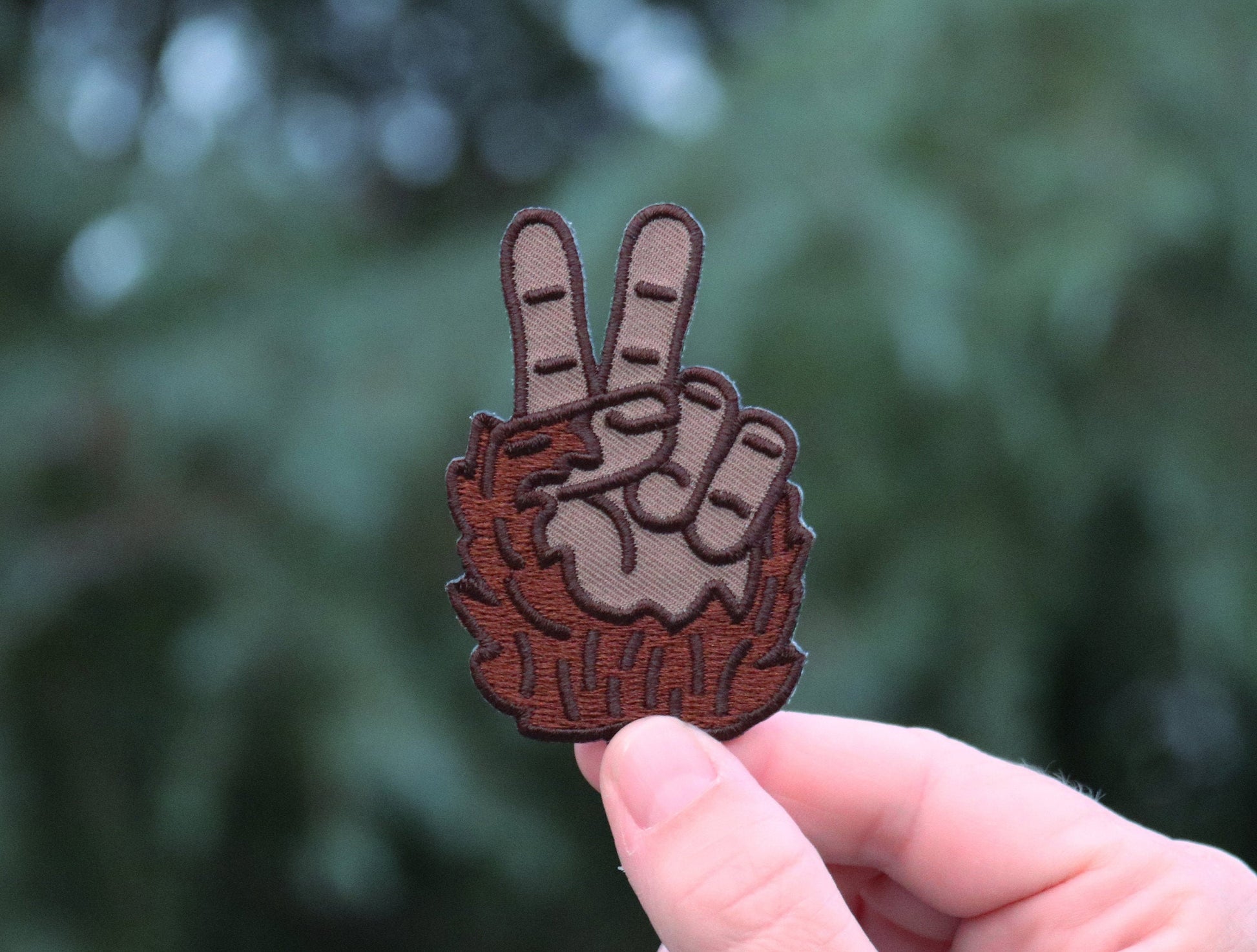 Held closer to the camera, the Bigfoot Peace Patch fills the frame with dark green trees behind it.