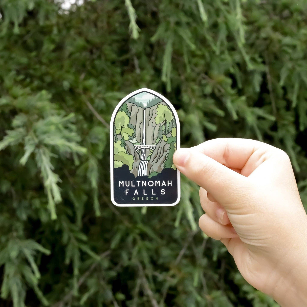 A hand holds the arched Multnomah Falls Oregon sticker in front of green conifer branches.
