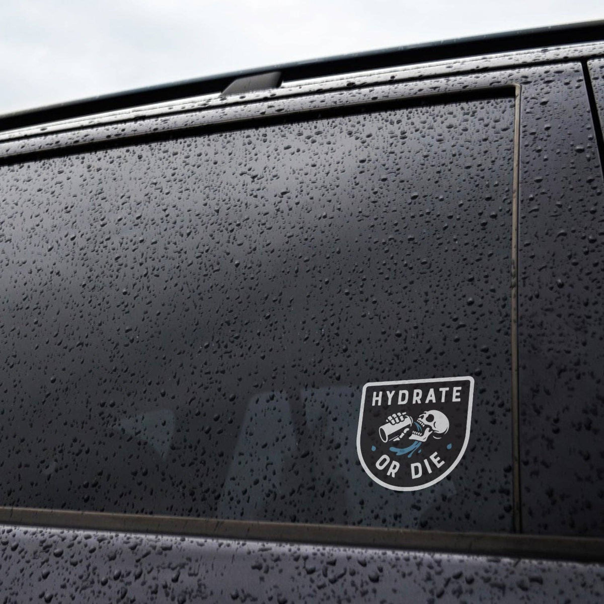 Rain beads across a dark car window where the Hydrate or Die sticker is applied to the glass.