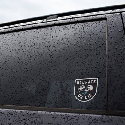 Rain beads across a dark car window where the Hydrate or Die sticker is applied to the glass.