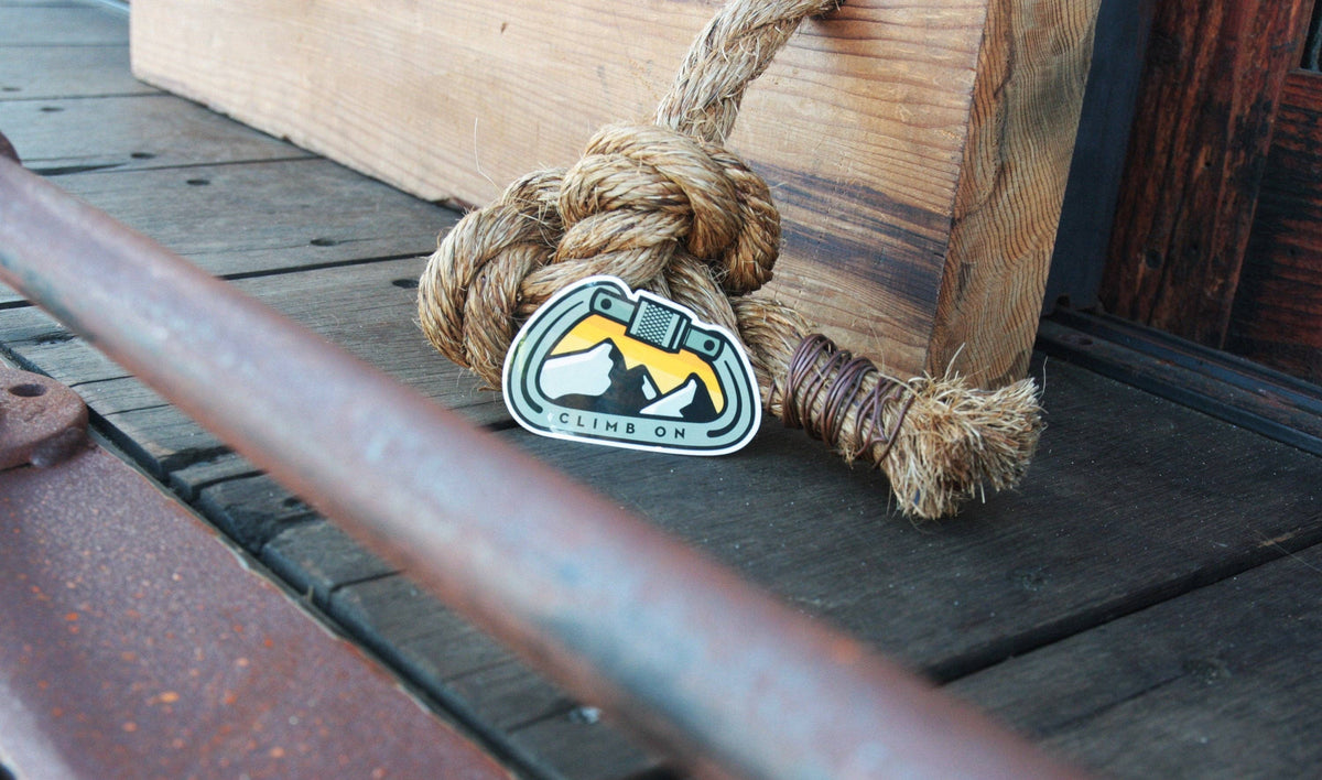 Propped against a knotted rope and timber, the Climb On Carabiner sticker rests on old dock boards.